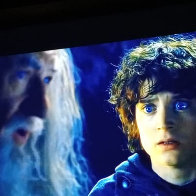 The image shows a close-up of two characters from the Lord of the Rings franchise, Gandalf and Frodo Baggins, with their eyes glowing bright blue. Gandalf, on the left, has a long white beard and appears to be speaking, with his mouth open. Frodo, on the right, has curly brown hair and looks startled, with his eyes wide open and focused on something off-camera. The background is a dark blue, creating a mystical and dramatic atmosphere. The lighting is focused on the faces of the characters, making their glowing eyes the most prominent feature. There is no information about the city of Zam'yany, Russia, in this image.