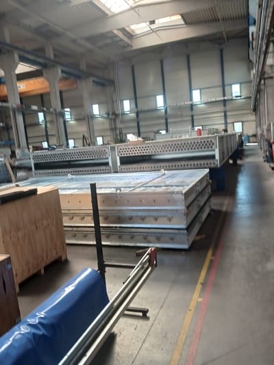 The image shows the interior of a large industrial facility, likely a factory or warehouse, in Benešov nad Ploučnicí, Czechia. The perspective is tilted, looking upwards and along the length of the building. The setting is a well-lit, open space with high ceilings supported by blue structural beams. Natural light enters through rows of rectangular windows high up on the walls.
The floor is concrete, marked with red and yellow lines, suggesting designated pathways or safety zones. Several large, metallic objects are visible, appearing to be stacks of industrial components or equipment. These are silver-colored and have a grid-like texture or perforation. One prominent object is a long, narrow, perforated metallic structure. Another set of objects are larger, box-like metallic structures, stacked in a few rows.
In the foreground, to the right, is a large roll of blue material, possibly a tarp or protective covering, resting on a wooden crate. The overall impression is one of an active industrial environment, with materials and equipment stored or in the process of being handled. There are no people visible in the frame. The time of day is not discernible, but the artificial and natural lighting suggest it is daytime. The weather is not relevant to the interior scene. No visible text is present.