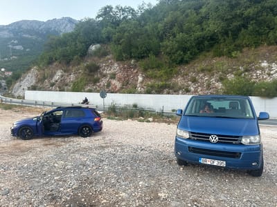 A blue Volkswagen van and a blue Volkswagen hatchback are parked on a gravel lot beside a road. The hatchback's driver's side door is open, and the interior is visible. The van has its headlights on and a person can be seen in the driver's seat. The license plate on the van reads "MNE BR DF 395". In the background, a mountainous, forested landscape is visible, with a concrete barrier running alongside the road. The sky is overcast, suggesting it is either early morning or late evening. The overall scene appears to be a rest stop or parking area in a rural, mountainous region.
