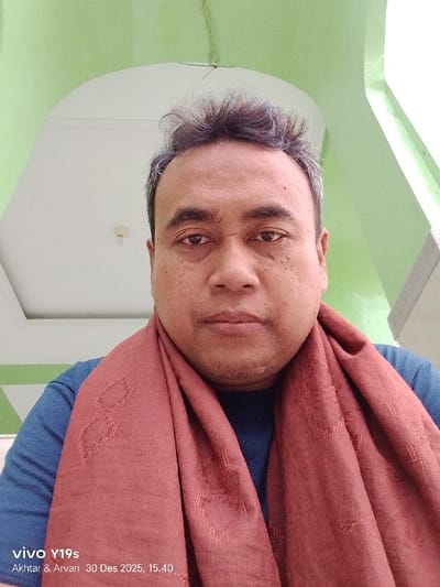 A close-up image, captured in Jagapura Lor, Indonesia, features an adult male positioned centrally and facing directly forward. The individual has dark hair with visible grey strands, a medium skin tone exhibiting minor facial irregularities, and dark eyes with a neutral expression. He wears a dark blue garment and a rust-colored scarf draped around his neck, displaying woven geometric patterns. The background consists of light green and off-white architectural surfaces, including a visible arched ceiling section on the left with a bare lightbulb. A watermark in the lower-left corner indicates "vivo Y19s Akhtar & Arvan 30 Des 2025, 15.40."