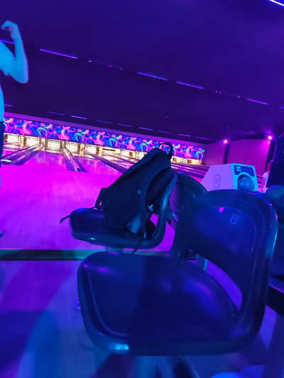 The image captures a bowling alley with a vibrant, neon-lit atmosphere, illuminated by purple and pink lighting. In the foreground, dark blue bowling alley chairs are prominent, with a black backpack resting on one of them. A portion of a person's arm and shoulder is visible on the far left, seemingly in motion. The bowling lanes stretch into the background, adorned with colorful, abstract patterns on the walls above. Two bowling balls are visible on a rack in the background, and a scoring console sits on a platform. The overall scene suggests a recreational setting, likely a bowling alley. However, there is no information to place this specific scene in Chelles, France.