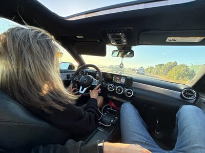 A woman with blond hair is driving a Mercedes-Benz, with the sunroof open, on a highway during daylight. The passenger, presumably a man, is wearing blue jeans. The vehicle is positioned in a lane with other cars, with green vegetation lining the side of the road. The interior of the car shows the dashboard and the rearview mirror, along with a hanging decorative charm. Based on the environment and road conditions, it is plausible the location is Huntington Beach, United States.