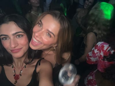 This is a medium-shot, selfie-style photograph taken in what appears to be a nightclub in Monteclaro-la Cabaña, Spain. Two young women are prominently featured in the foreground, looking towards the camera and smiling. The woman on the left has dark hair and is wearing a black top with a beaded necklace. The woman on the right has lighter brown hair and is wearing large, gold-colored earrings. Both appear to be enjoying themselves. In the background, other people and green lighting are visible, suggesting a lively atmosphere. One person in the background appears to be holding a glass. The overall impression is one of a celebratory night out.