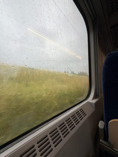This media file shows a view from inside a train compartment looking out a rain-streaked window. The window is covered in numerous water droplets, obscuring the view of the outside landscape. The outside appears to be a mix of overcast sky and a greenish-brown field, suggesting a rural or semi-rural setting. A faint, striped reflection is visible on the left side of the window, possibly from the train's interior.

The interior of the train compartment is partially visible. In the upper part of the image, a blue patterned seat is seen, along with some overhead luggage racks. To the right of the window, a beige-colored vent with horizontal slits is present. The overall impression is a dreary, rainy day, likely in the daytime, with the view from the train conveying a sense of travel. No people are visible in the frame. There is no visible text. The location context of Żurawica, Poland, indicates a travel scenario within that region.