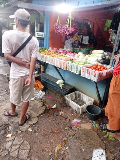 The image shows a street vendor stand with various produce on display in Kecamatan Caringin, Indonesia. The stand is located at the coordinates latitude: -7.523, longitude: 107.485.  There is a man wearing a white shirt, khaki shorts, and a white baseball cap standing in front of the stand. He is looking at the produce. There are also various other fruits and vegetables on display, including bunches of grapes, mangoes, oranges, and tomatoes.  The stand is made of wood and has a blue and white painted surface. The scene is set in an urban environment with a stone and dirt walkway in front of the stand. The ground is covered with debris and leaves. There is an empty plastic crate on the ground next to a black plastic bucket. A second person, partially visible, wearing orange pants is standing on the other side of the stand, facing the opposite direction of the first person.