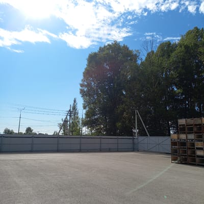 A sunlit, wide-angle shot captures an outdoor industrial or storage area in Kinel, Russia, on a clear day. The foreground is dominated by a vast expanse of asphalt, reflecting a pale green sun glare diagonally from the lower right to the mid-left. A long, corrugated metal fence, approximately ten feet high, stretches across the middle ground, creating a visual division. Behind the fence, a dense line of lush green trees forms a natural backdrop. A tall, weathered metal gate with a geometric pattern is visible on the right side of the fence. To the far right, a towering stack of open-fronted metal containers, possibly for storage or transport, rises several stories high. In the sky, a bright sun shines intensely in the upper left, casting long rays through scattered white clouds. Power lines and poles are visible in the upper left portion of the image, extending horizontally across the frame. The overall scene suggests an operational but possibly underutilized industrial or logistics facility.
