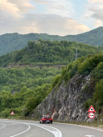 A landscape photograph shows a mountainside road in Očinići, Montenegro. The road curves sharply. A red car is visible on the road. Road signs indicate a sharp turn and restrictions on vehicle width. The mountain is covered in dense green vegetation. A transmission tower is positioned on the mountainside. The sky is overcast. The photograph is oriented vertically.