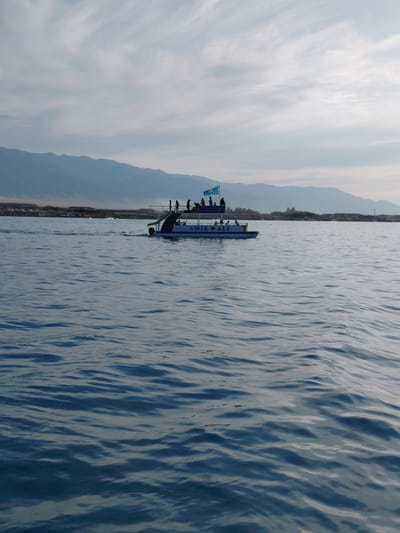 This is a wide-angle, landscape orientation photograph taken from a body of water, likely a lake or sea, showing a boat with people on it and a distant shoreline with mountains. The image is rotated 90 degrees clockwise, so the horizon appears vertical.
The foreground is dominated by the rippling blue water. On the left side of the image, a boat is visible, traveling parallel to the distant shore. Several people are standing on the deck of the boat, some are silhouetted against the sky. A flag, which appears to be the flag of Kazakhstan, is flying from the boat. The text "MIR ALI" is visible on the side of the boat, with a heart symbol between the two words.
In the background, a low shoreline is visible, dotted with buildings and trees, suggesting a populated area. Beyond the shoreline, a range of mountains stretches across the frame, their peaks softened by distance and the atmospheric conditions. The sky above is cloudy, with streaks of light suggesting it is either early morning or late afternoon. The overall mood of the image is serene and peaceful, with the vastness of the water and mountains creating a sense of calm. The location cues, such as the flag and the mountainous landscape, suggest the scene is in Akshi, Kazakhstan.