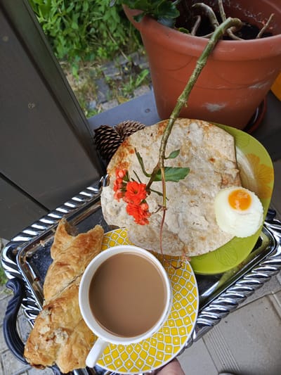 A close-up, high-angle shot shows a breakfast tray on a dark wooden surface. The tray contains a white mug with a brown beverage, a pastry, a green plate with a flatbread and a poached egg, and two pinecones. A plant with orange flowers and green leaves arches over the flatbread from the direction of a terra cotta pot. The background shows greenery and the edge of a building. The location is Alexandrovskaya, Russia.