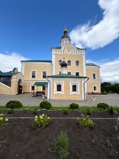 The image depicts a religious building, likely a church or monastery, with a pale yellow facade and a dark green roof. The building features multiple domes, one of which is topped with a golden spire. The architecture is ornate, with decorative window surrounds and cornices. In the foreground, a landscaped garden bed filled with dark soil contains several small green bushes and some flowering plants with pale yellow blossoms. A paved walkway with a brick border separates the garden from the building's courtyard, which is also paved with interlocking stones. The sky above is blue with scattered white clouds, and a small bird is visible in flight. The building appears to be situated in an urban or semi-urban environment, with hints of greenery and trees visible in the background. The overall scene conveys a sense of peace and architectural beauty. This specific structure is the Resurrection Cathedral in the city of Lubny, Russia.