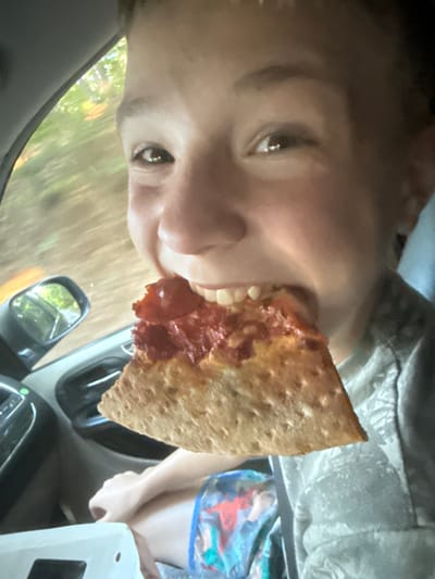 A close-up, indoor, eye-level shot shows a young person in the driver's seat of a vehicle, biting into a slice of pizza. The person's face is the primary focus, with their mouth open as they take a bite. The pizza slice is a triangle with a golden-brown crust and red sauce and pepperoni. The background shows the interior of the vehicle, including the dashboard and a side mirror reflecting a blurry green and brown outdoor scene. The lighting is natural and appears to be daylight. The image was taken in Emerson, United States of America.