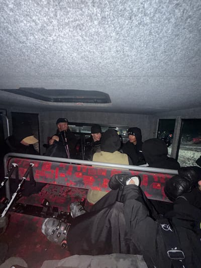 The image shows the interior of a bus in L'Alpe d'Huez, France. Several people are seated, mostly dressed in dark clothing. One person is wearing ski boots and holding ski poles. The bus seats are red with a repeating building pattern. The walls are gray carpeted. Outside the window, it appears to be nighttime or low light conditions.