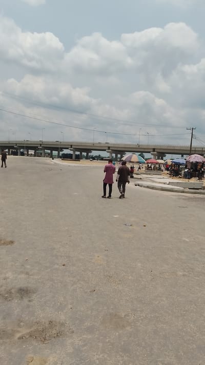 The image captures a daytime scene in Effurun, Nigeria, featuring a wide, unfinished concrete road under a cloudy sky. An elevated highway bridge spans the background. Two men walk away from the viewer on the road, one wearing a magenta shirt, the other in a brown shirt. A smaller figure is visible walking on the far left. To the right, under the bridge, there are small vendor stalls with colorful umbrellas and people gathered around. A power pole is visible near the stalls. The road surface shows patches of discoloration and unevenness.