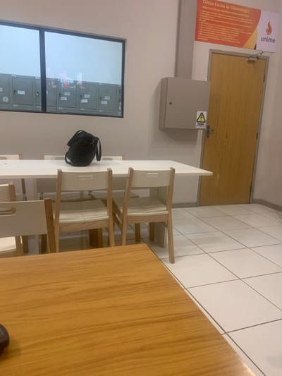 The image depicts an unoccupied interior room at the "Clínica Escola de Odontologia" (Dental School Clinic) within the Unime university campus, located in Lauro de Freitas, Brazil. The environment features light-colored walls and a white tiled floor. In the mid-ground, a long white rectangular table is surrounded by multiple light-wood chairs with white seats and backrests. A black shoulder bag rests centrally on this table. To the left, a large horizontal window offers a partial view of what appears to be a row of gray lockers or storage units. To the right, a light-colored wooden door is visible, positioned beneath a red and orange sign bearing the text "Clínica Escola de Odontologia" and the "unime" logo. Adjacent to the door, a gray electrical box is mounted on the wall, displaying a yellow triangular "CUIDADO" (Caution) sign with a lightning bolt symbol. A portion of a brown wooden table is visible in the lower foreground. No individuals are present in the scene.