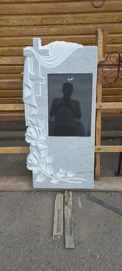 A grey granite headstone with a dark granite inset panel is displayed outdoors. The headstone features an embossed carving of a draped cross and roses on its left side. A silhouetted figure of a person, likely the photographer, is reflected in the dark panel, holding a mobile phone. The headstone is positioned in front of a wooden slatted wall and resting on wooden planks, on a gravelly surface. The overall scene suggests a workshop or display area for funerary monuments, likely in Ertis, Kazakhstan, though this specific location is not visually confirmed.