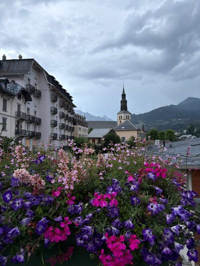 The image captures a view of Saint-Gervais-les-Bains, France, focusing on a foreground of vibrant pink and purple flowers, likely petunias and geraniums, in a planter. Behind the flowers, a light-colored building with multiple balconies and a dark roofline dominates the left side of the frame. To the right of this building, a church steeple rises above other structures, with the silhouettes of mountains visible in the background under a cloudy sky. The scene suggests a town nestled in a mountainous region, with floral displays contributing to the urban aesthetic. There are no people visible in the image.