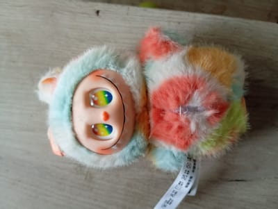 The image shows a plush toy lying on a wooden surface. The toy is oriented on its side, with its head facing left. It has a round, light-skinned face with large, expressive eyes that feature rainbow-colored irises. A small, curved black line represents its mouth. The toy is wearing a fluffy, multi-colored hooded garment. The hood is predominantly light blue with some orange accents on the ears. The body of the toy is covered in soft, fluffy material in a tie-dye pattern of pink, orange, yellow, and green. A white tag is attached to the toy, featuring various symbols and text in what appears to be Chinese. The wooden surface beneath the toy has a visible grain pattern. The lighting suggests an indoor setting, likely during the daytime, with no specific weather cues. The overall impression is of a cute and whimsical toy.