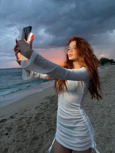 A woman with long, red hair stands on a beach, holding a small, black device emitting a bright light. She is wearing a sheer white dress with long sleeves. The beach consists of sand and small pebbles, with waves visible in the background. The sky is dark and cloudy, suggesting either dawn or dusk. The location is Alimos, Greece, based on the beach and landscape characteristics.