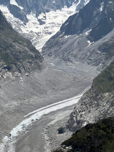 The image depicts a wide, rugged glacial valley in Chamonix-Mont-Blanc, France. Steep, rocky mountain slopes covered with scree and sparse vegetation flank the valley. A long, winding glacier, appearing white and icy, flows down the center of the valley, interspersed with rocky debris. The upper reaches of the valley are dominated by extensive snowfields and glaciers clinging to the higher mountain peaks. The overall scene is one of a raw, natural mountain environment, likely taken from a vantage point overlooking the glacial expanse.