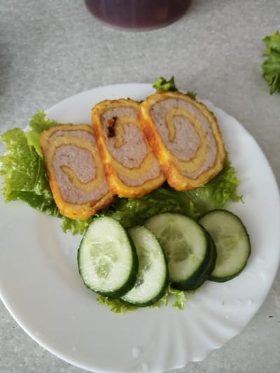 A white plate holds three rolled food items on a bed of lettuce. Each roll features a spiral of light brown meat and a yellow outer layer, possibly egg or pastry. Sliced cucumbers are arranged in front of the rolls. The plate sits on a speckled gray surface. In the upper background, a dark, out-of-focus object, likely a drink in a glass, is visible. A sprig of lettuce is also visible to the right of the plate. This scene is not located in Bezlesnyy, Russia.