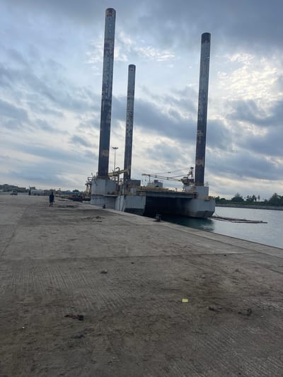 The image shows a port scene in Ebute Leki, Nigeria. Three large, cylindrical metal towers rise vertically from a platform attached to a dark-colored barge or ship docked alongside a concrete pier. Cranes and other machinery are visible on the platform. The sky is cloudy. A person is walking in the distance along what appears to be a road or walkway parallel to the pier. Concrete structures are on the far right, and some trees are across the water in the distance.