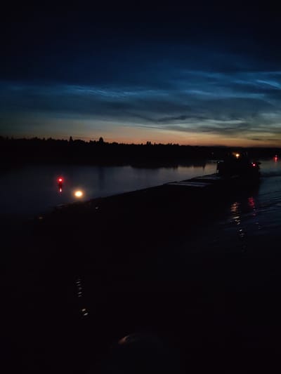A nighttime view of a barge moving on a body of water, possibly a river or canal, near the city of Kovrizhnovo, Russia. The sky displays a gradient of dark blue at the top transitioning to lighter hues of orange and yellow near the horizon, with wispy clouds illuminated by the fading light. Silhouetted trees line the far bank, creating a dark, indistinct treeline. In the foreground, a barge is partially visible, with its deck appearing dark and wet. A single red navigation light is prominent on a post near the water's edge, with a brighter, yellowish light from the barge itself. The water reflects some of the ambient light and shows ripples. The scene is characterized by low light conditions and a sense of calm.