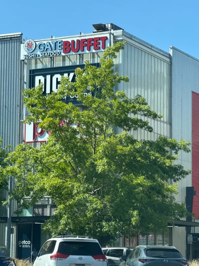 The image depicts a commercial building on a clear, sunny day in Seattle, Washington, USA. The building features modern architecture with corrugated metal siding and large glass panels. Prominently displayed on the upper facade are signs for "GATE BUFFET SUSHI SEAFOOD" and "DICK'S." Below, at street level, a sign for "petco THE HEALTH + WELLNESS CO." is visible. A lush green tree with dense foliage partially obscures the building's lower sections. Several vehicles are parked or driving in front of the building, including a white Toyota Sienna minivan and a dark grey Mazda CX-5. The overall scene suggests a commercial district with various businesses.