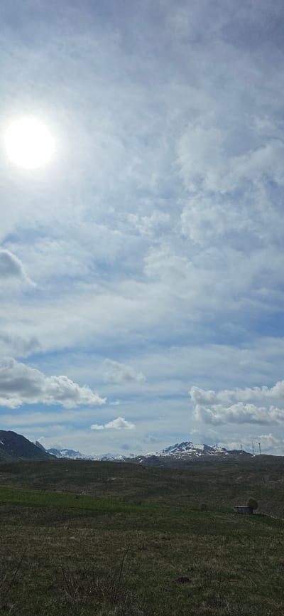 The image depicts a landscape in Krnovo, Montenegro, showing a grassy hillside under a partially cloudy sky. In the foreground, there is grass and some small plants. Snow-capped mountains are visible in the distance. Several wind turbines are visible on the hill. The sky is blue with white clouds, and the sun is bright.