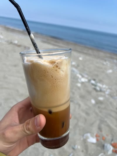 A hand holds a glass of iced coffee with a black straw, likely a frappe or similar blended coffee drink, with a thick layer of foam at the top. The background reveals a beach with pale sand and the Aegean Sea under a bright, overcast sky. The image likely depicts a person enjoying a coffee at Leptokarya Beach in Leptokarya, Greece. The focus is on the coffee and the beach setting, with the human element serving as a point of perspective. The scene is peaceful, with the gentle tones of the sea and the refreshing nature of the beverage suggesting a relaxed atmosphere.