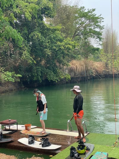 Two individuals are on a wooden dock at a lake, likely preparing for water sports. A man in a black long-sleeved shirt, orange shorts, and a sun hat stands near the water's edge, with a metal ladder extending into the water. Next to him is a wakeboard and bindings. A younger person with dyed blue hair, wearing a white t-shirt and blue shorts, stands further back on the dock. A life vest lies next to the individual. Various wakeboards and accessories, including bindings and an orange bucket, are also on the dock. The background consists of lush green trees and reeds growing along the edge of the lake. The location is Tambon Nam Phrae, Thailand.