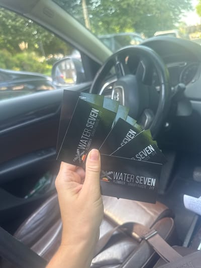 The scene is an interior shot of a car, presumably in Marcq-en-Barœul, France, based on the business cards held by a hand in the foreground. The hand holds several business cards for "WATER SEVEN," a plumbing and heating company ("PLOMBIER CHAUFFAGISTE 7/7"). The car's steering wheel and dashboard are visible in the background, suggesting the photo was taken from the driver's seat. The car seat and seatbelt are also visible. Outdoor bokeh light spots suggests the presence of greenery.