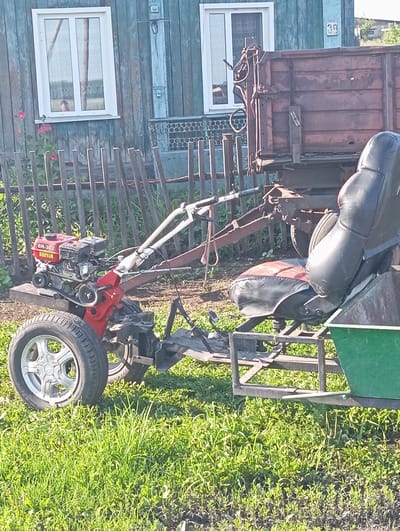 In Новокопылово, Russia, a red and silver walk-behind tractor is parked on a grassy area in front of a weathered blue wooden house. The tractor has a single wheel on the left side, a red engine with "BR-302" visible, and a black seat with red accents attached to a metal frame extending from the tractor. Behind the tractor, a rusty brown trailer with wooden planks is hitched. The house has two white-framed windows, and a wooden fence is situated in front of it. The environment is rural, with lush green grass and some plants visible in the foreground. The sky is bright, suggesting daylight.