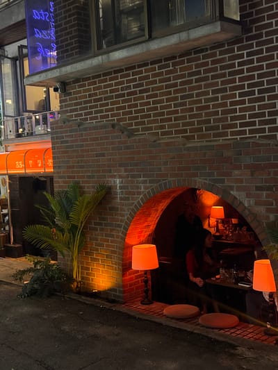 The image depicts a nighttime street scene in 마포구, South Korea, featuring the exterior of a dark red brick building. A large, deeply recessed brick archway at ground level serves as a dimly lit entrance or outdoor seating area. Inside the archway, warm, orange-toned light illuminates a bar or counter setup where bottles and glassware are visible. One female individual with dark hair is partially visible, seated at this counter. Another person is partially obscured in the shadows further inside the archway.
Two identical floor lamps with orange fabric shades stand on the pavement, flanking the archway's opening. Two circular, orange floor cushions are placed directly in front of the archway on the dark asphalt pavement. To the left of the archway, a tall, artificial palm plant is positioned against the brick wall, accompanied by smaller green foliage at its base.
Above this plant, an orange retractable awning displaying partial Korean characters, "53 - Yu K", extends from an adjacent section of the building. Further left and higher, a windowed upper floor exhibits a blue neon sign with stylized or inverted text. A small surveillance camera is mounted on the brick wall below the windows. The foreground consists of dark asphalt pavement.