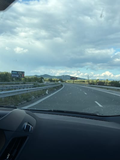 The image depicts a view from inside a vehicle traveling on a highway near Blajievo, Bulgaria. The road is a multi-lane highway with guardrails. The sky is cloudy with some patches of blue. Trees and vegetation line the side of the road. A billboard is visible on the side of the highway, displaying an advertisement with numbers and text. The dashboard and windshield of the car occupy a significant portion of the foreground.
