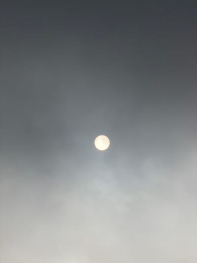 The image depicts the sun, partially obscured by clouds, in the sky above Ngadas, Indonesia. The sun appears as a bright, circular orb with a yellowish-white hue, and its light diffuses through the overcast atmosphere, creating a hazy effect. The sky transitions from a darker gray at the top of the frame to a lighter, more muted gray towards the bottom. There are no visible people, buildings, or other distinct objects in the frame, just the sky and the sun. The overall impression is one of a subdued, atmospheric condition in the sky.