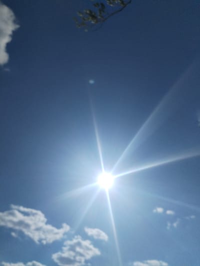 A bright sun with visible rays dominates the center of the image, positioned against a clear blue sky. Several white, puffy clouds are scattered across the lower portion of the frame, with some appearing blurred due to motion or focus. A small branch with green leaves is visible in the upper right corner, slightly out of focus. The overall impression is of a sunny day. There are no people or animals visible in the image. The image does not contain any recognizable landmarks or features specific to Bilousivka, Ukraine.