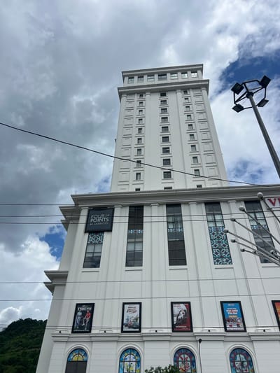 The image shows a tall, white, modern building in Hà Giang 2 Ward, Vietnam, under a cloudy sky. The building has many windows arranged in a grid pattern. A prominent sign for "Four Points by Sheraton" is visible on the building. Several framed advertisements or posters are also displayed on the lower facade of the building. A street light fixture with multiple lamps is positioned to the right of the building. A lush green hillside is visible in the background to the left. Electrical wires crisscross the scene in the foreground.