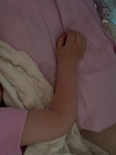 This media file shows a close-up of a child's arm resting on a pink blanket. The child is wearing a pink garment. A white fluffy blanket is partially visible. In the background, a light blue object with a cartoon cat face can be seen. The scene appears to be indoors, possibly in a bedroom, given the blankets. The lighting suggests it might be daytime or a well-lit room. The overall impression is one of comfort and rest. There is no visible text in the image.