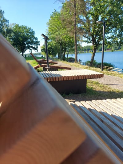 The image depicts a waterside park with modern wooden benches and lampposts. A tranquil blue lake stretches across the background, bordered by lush green trees and a clear blue sky. In the foreground, the close-up view of a wooden bench draws attention to its slatted design and warm wood tones. The park setting suggests a recreational area, possibly in the city of Razvetye, Russia, given the provided context. The overall atmosphere is peaceful and serene, with the sunlight casting gentle shadows across the scene. No people are visible in the image, emphasizing the quiet and natural beauty of the location.