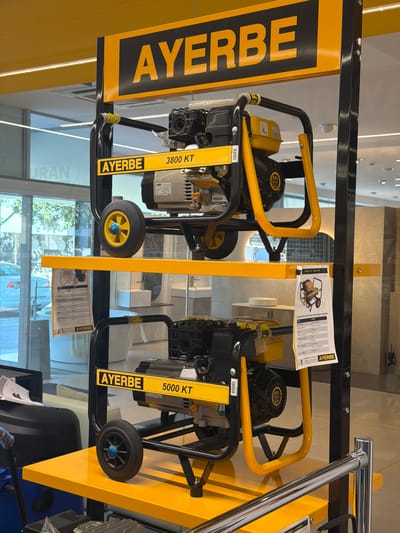 This media file shows a retail display of two yellow and black generators, branded "AYERBE". The generators are positioned side-by-side on a black metal frame, with yellow accents. Both generators have black wheels, yellow handles, and prominently displayed "AYERBE" branding. One generator has a yellow label with "3800 KT" and the other has "5000 KT". A technical specification sheet for the "AY-5000 KT MA" model is visible, along with other product information labels. The setting appears to be an indoor retail space, possibly a hardware or industrial equipment store, given the shelving and lighting visible in the background. There are no people in the image. The lighting suggests it is daytime. The text "URAN" is visible in the background, potentially indicating a location or another brand. The overall scene is a product showcase.
