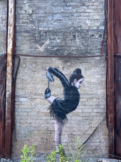 A mural depicts a young ballerina in a black leotard and tutu, performing a difficult pose against a weathered brick wall. The ballerina is on her tiptoes, with her leg extended upwards and bent backwards, her arms reaching up to grasp her foot. Her hair is pulled back in a bun. The mural is located in Horenka, Ukraine. The brick wall shows signs of age and decay, with cracks and discoloration. Two rusted metal beams frame the sides of the mural, suggesting it may be on the exterior of a damaged or abandoned building. In the foreground, green plants are growing, indicating an outdoor setting.