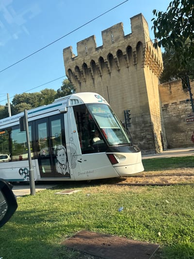 The image captures a modern white tram with a portrait of Daniel Auteuil on its side, stopped near a section of the ancient city walls of Avignon, France. The tram is on a track that runs alongside a grassy area. In the background, the imposing stone ramparts of the city walls, characterized by crenellations and arched openings, are prominently visible against a clear blue sky. The overall scene depicts the juxtaposition of contemporary urban transit against historical architecture on a sunny day.