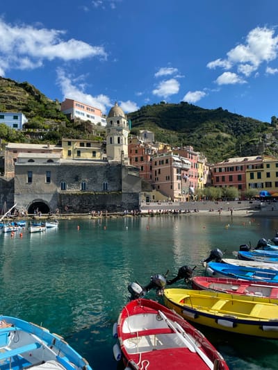 The image displays a coastal scene in Vernazza, Italy, centered on its harbor under a bright blue sky with sparse cumulus clouds. In the immediate foreground, a line of small, multi-colored boats (red, yellow, blue, and white) equipped with outboard motors and some with oars, are moored in the clear, emerald-green water. Orange and white buoys are visible in the water.
The midground is dominated by the Church of Santa Margherita d'Antiochia, a stone-grey edifice featuring a large arched opening at its base and arched windows. Adjacent to the church's right, a cream-colored bell tower with a domed cupola and a visible clock face ascends. The town's architecture, characterized by multi-story buildings painted in shades of terracotta, yellow, pink, and light green, spreads around the harbor and climbs the steep, terraced, green hillsides in the background.
The shoreline is populated with numerous individuals. Groups are observed sitting on the rocky outcrop to the left of the church's archway, and others are distributed across the sandy beach area to the right, some standing, some seated. The environment suggests a sunny, calm day.