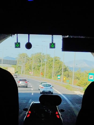 The image shows a view from inside a vehicle exiting a tunnel onto a highway. The time of day appears to be daytime, with bright sunlight visible. Two green arrows pointing downwards are displayed on a gantry above the highway, indicating the direction of travel. A large, dark, circular object, possibly a light or a sign, is also present on the gantry to the right of the arrows.

Several cars are on the highway, including a gray hatchback in the left lane and a white hatchback in the right lane. A truck with a white trailer is visible in the distance on the left side of the highway. Road signs and streetlights line the highway.

The landscape beyond the highway includes green trees and hills in the distance, with a body of water and mountains visible under a clear blue sky. The right edge of the image shows the silhouette of a person or object in the foreground, obscuring part of the view.

The presence of signs indicating a highway, traffic signals, and road infrastructure suggests this is a public road. The overall scene depicts normal traffic flow on a sunny day. The Municipal Unit of Ampelakia is located in the region of Attica, Greece, and this highway is likely part of the Greek national road network.