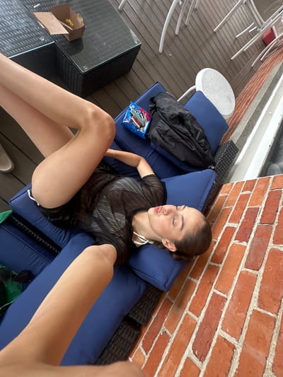 A young woman is lying on a blue lounge chair on a wooden deck next to a brick wall. She is wearing a sheer black top and black shorts, and her legs are raised in the air. Her eyes are closed and her lips are puckered. A black jacket and a bag of chips are on the lounge chair next to her. A black table with a takeout container on it is to the left of the lounge chair. The scene appears to be an outdoor patio in Harbor Springs, Michigan.
