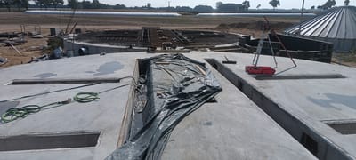 This is a medium-angle, eye-level, exterior, full shot of a construction site in Magnuszewice, Poland. The foreground is dominated by a large, gray, concrete surface, featuring several rectangular openings. A green garden hose is coiled on the concrete, with one end running towards the openings. A large section of the concrete surface is covered by dark gray plastic sheeting, which appears to be protecting something beneath it. To the right, a red tool carrier sits on the concrete, next to a tripod with surveying equipment. In the midground, construction materials including rebar and wooden forms are visible, surrounding a circular concrete structure with rebar reinforcement. In the background, a large, silver, conical silo stands next to a white, tarp-covered structure, possibly agricultural feed storage, in a field. Trees and utility poles are visible on the horizon. The scene suggests ongoing construction or preparation for future development.
