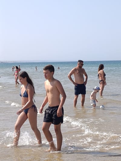 This is a photograph taken at a beach in Sines, Portugal. The scene depicts several people enjoying the ocean on a sunny day, likely in the afternoon, given the bright light and clear sky. The water is relatively calm with small waves breaking on the shore.

In the foreground, a young woman and a teenage boy are walking through the shallow water. The woman is wearing a blue bikini, and the boy is wearing black swim shorts. Both appear to be casually enjoying their time at the beach.

Further in the background, a man in dark blue swim shorts and sunglasses stands with his hands on his hips, looking towards the water. Beside him, a young child in striped swim shorts and a white cap is playing with a white ball in the water. A woman in a black bikini stands to the right of the man.

Other people can be seen in the water and on the shore in the distance, suggesting a moderately busy beach day. The overall atmosphere seems relaxed and summery. The horizon shows a faint line of land in the distance, typical of a coastal view. There is no visible text in the image.