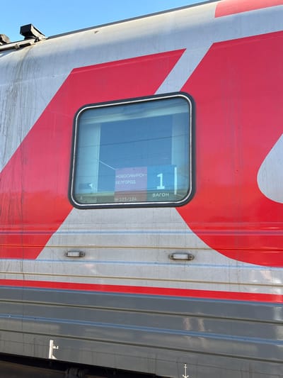 The image presents a forensic view of the exterior side of a passenger train car, stopped in Kropachyovo, Russia, under a clear blue sky. The upper section of the train car is silver-grey, adorned with large, stylized red branding, likely associated with Russian Railways (RZD). A dark grey lower section is separated by a thin red horizontal stripe. A rectangular window with rounded corners is visible, featuring an internal display. This display shows route information in Russian: "НОВОСИБИРСК-БЕЛГОРОД" (NOVOSIBIRSK-BELGOROD) as the destination, "1" as the wagon number, and "№ 123/124" as the train number, along with the word "ВАГОН" (VAGON). The train's exterior exhibits visible streaks of dirt and weathering, particularly on the silver-grey panels. Below the window, two small, rectangular light fixtures are embedded in the side. Technical markings are present on the dark grey lower panel. No individuals or discernible actions are observed in the scene.