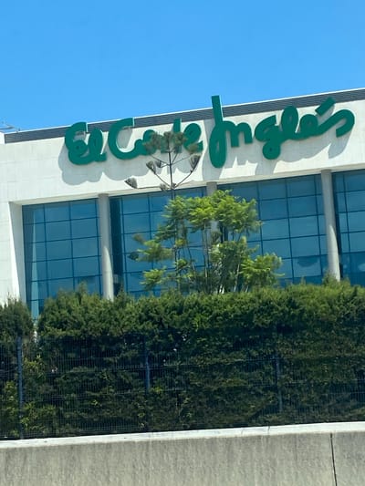 This image shows a close-up view of the "El Corte Inglés" department store facade in Nueva Andalucía, Spain. The store has a white exterior with a large, green, cursive logo spelling out "El Corte Inglés" prominently displayed at the top. Beneath the logo, large blue-tinted windows reflect the bright blue sky. In front of the building, a line of dense, dark green bushes forms a hedge, with a black metal fence visible behind it. A tall, slender tree with sparse, green foliage stands in front of the windows, partially obscuring the view of the building. The scene is set against a clear blue sky, suggesting a sunny day.