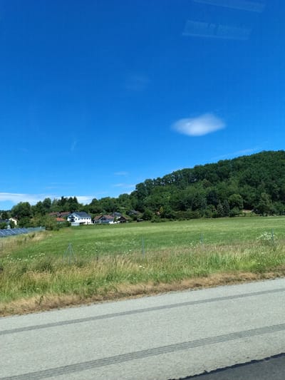 The image depicts a scenic view of a rural area with houses and a forested hillside under a clear blue sky. In the foreground, a paved road with tire marks runs horizontally, with a grassy area and a fence separating it from a green meadow. A small cluster of houses with red and white roofs is visible in the middle ground, nestled among trees. Solar panels are also present on the left side of the houses. The background is dominated by a dense green forest that covers a hillside. A single, small white cloud is visible in the sky. The overall impression is one of a peaceful, rural landscape. The location is identified as Steinach, Germany.